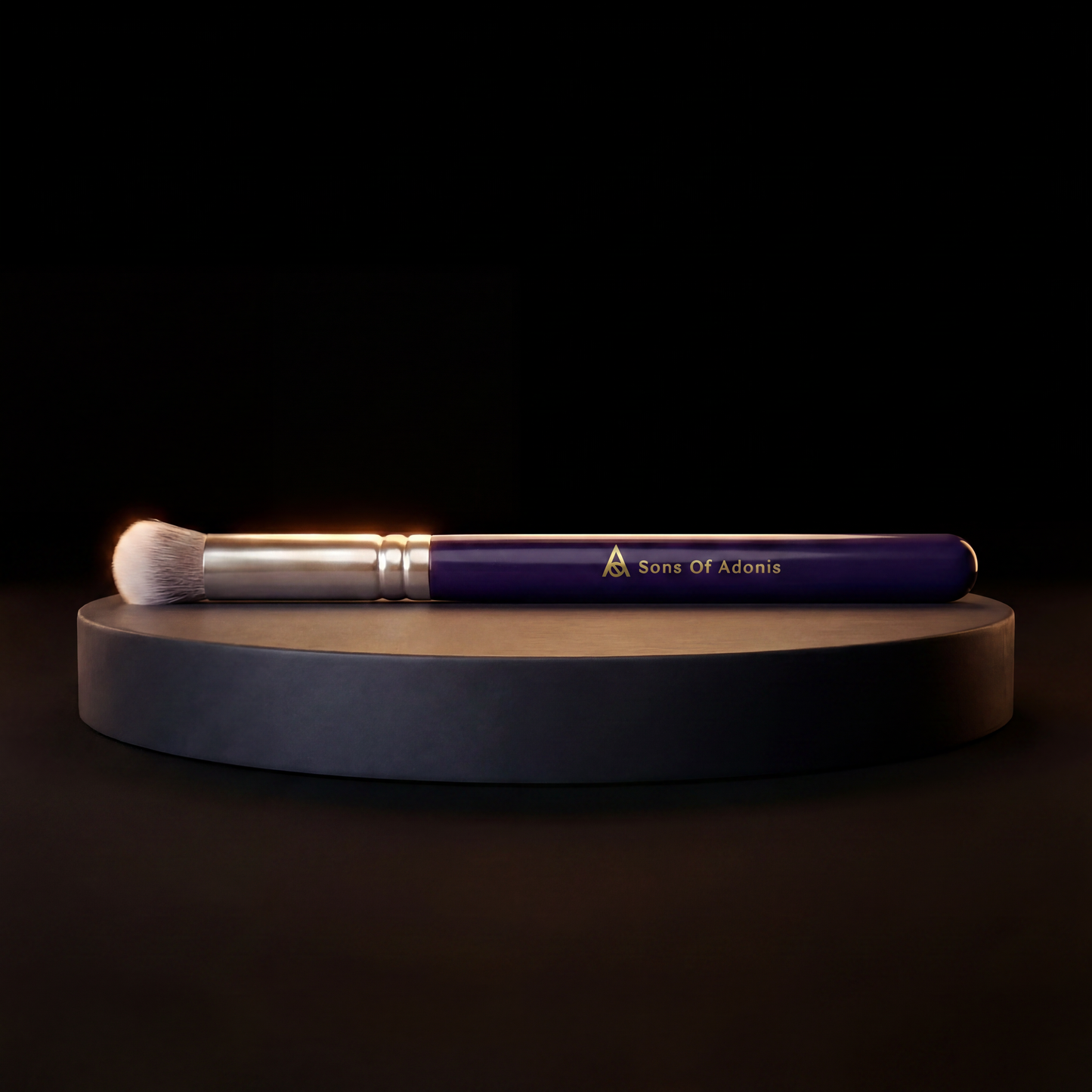 A makeup brush with a deep blue handle, silver ferrule, and white bristles, resting horizontally on a round black pedestal against a black background. The handle features the gold-foiled logo and text 'Sons Of Adonis'.